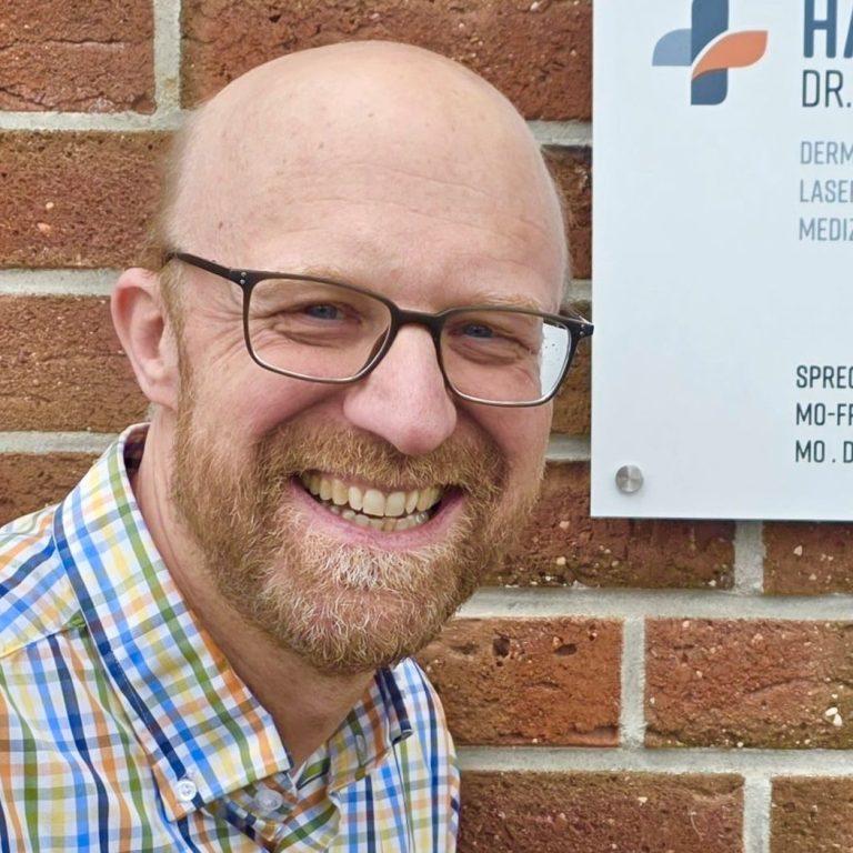 Dr. Alexander Dammann Mann mit Brille und kurzem Bart, trägt ein kariertes Hemd, lächelt vor moderner Architektur.